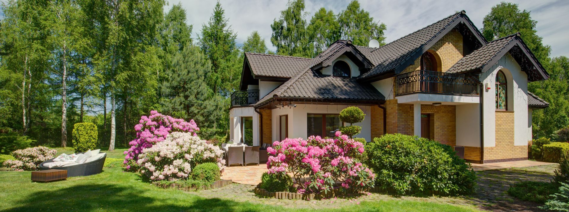 A house with a large yard, surrounded by trees and blooming flowers, on a sunny day.