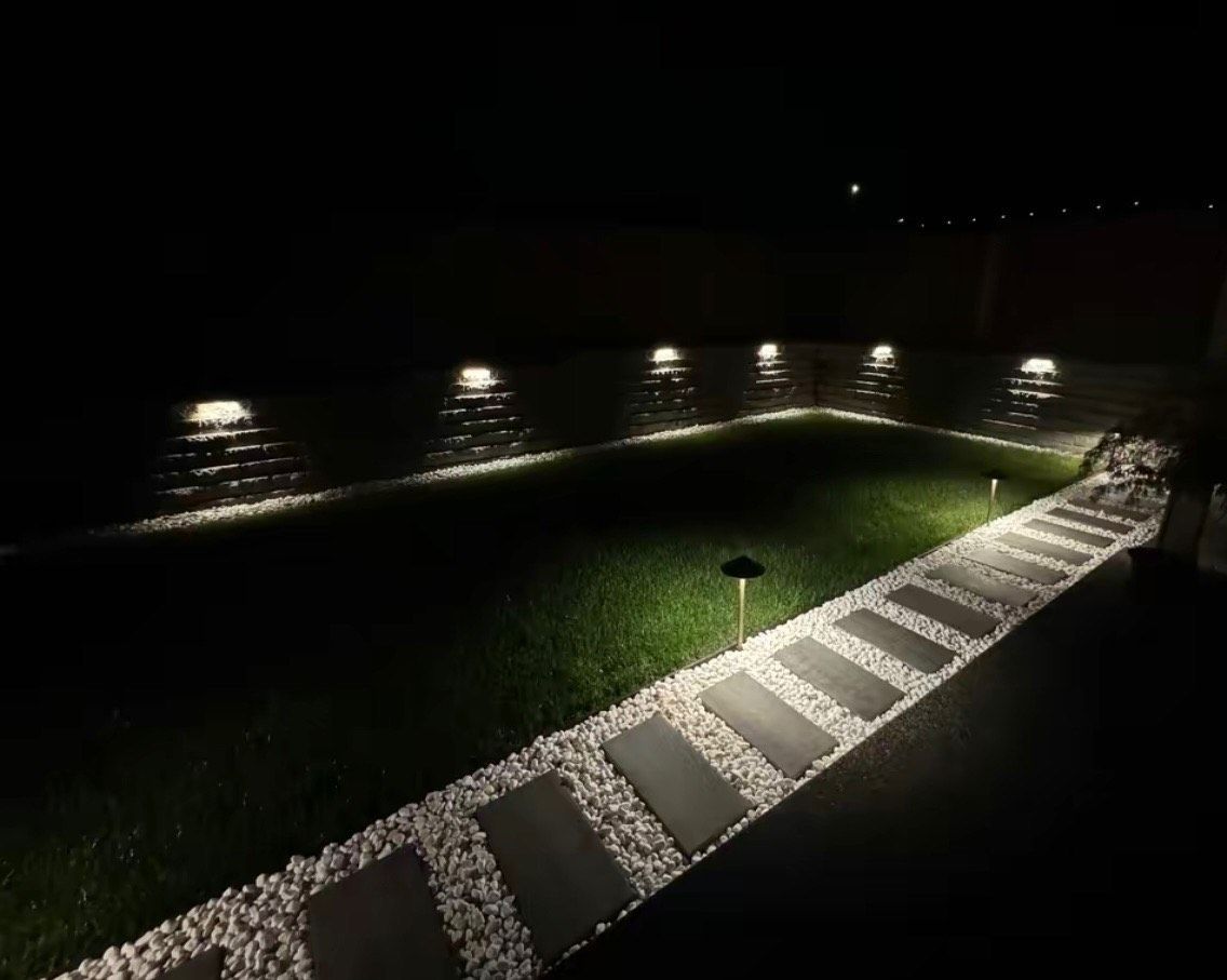 A lit garden at night with path lights illuminating plants, shrubs, and a grassy lawn.