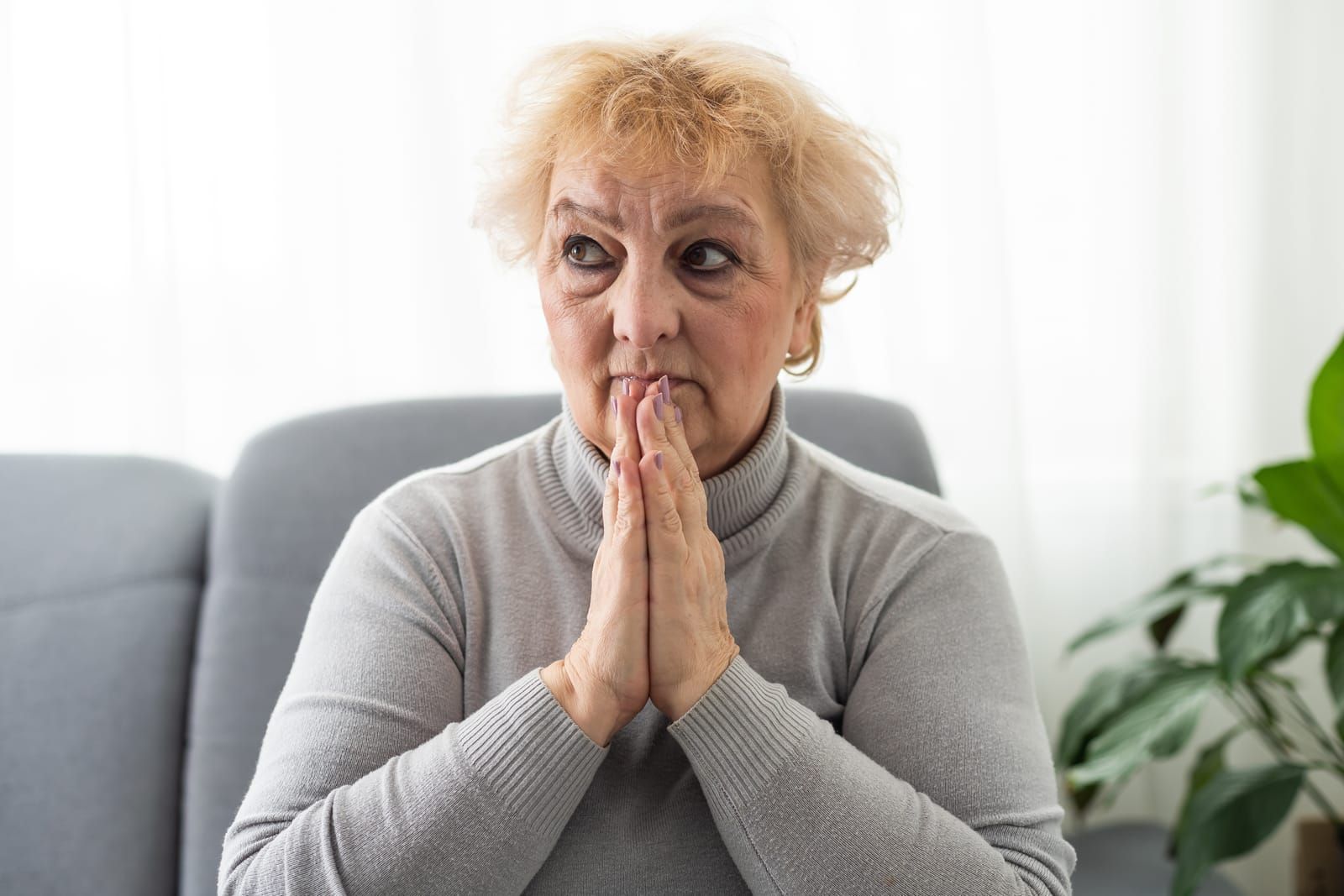 A person in a gray turtleneck sitting with their hands clasped together near their face, looking off to the side.