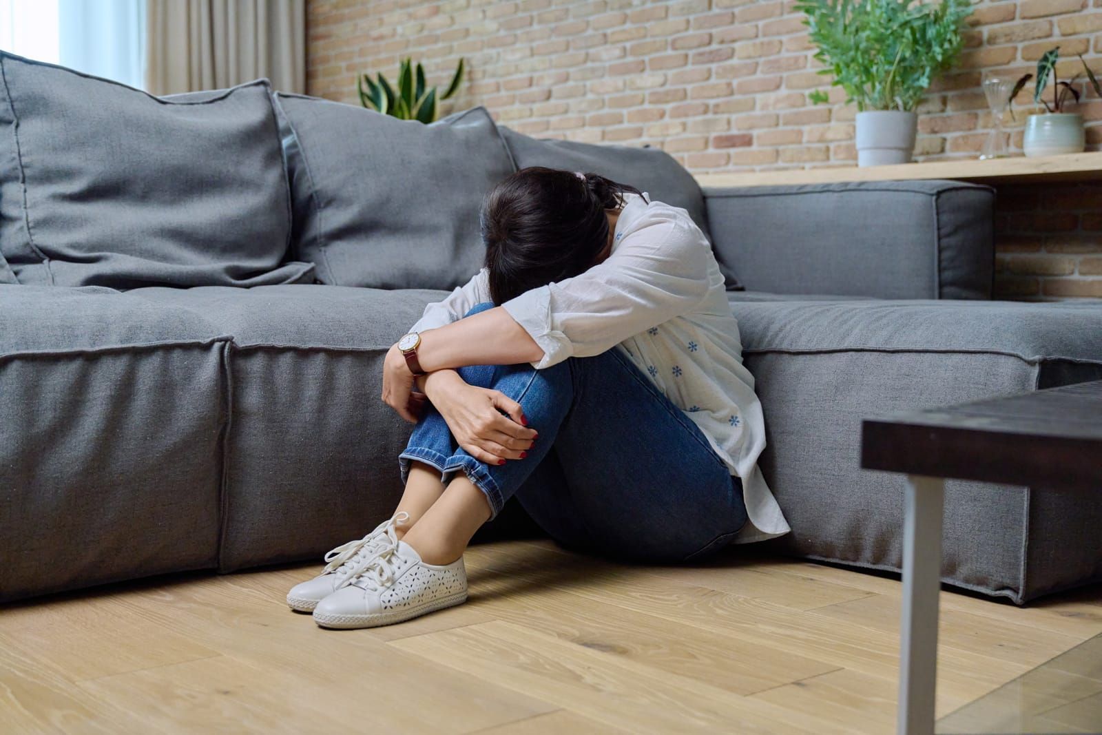 A person sits on a wooden floor, curled into a ball with their head down, next to a grey sofa in a living room.