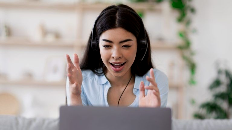Woman with headset using laptop, gesturing excitedly. Light blue shirt, home setting.