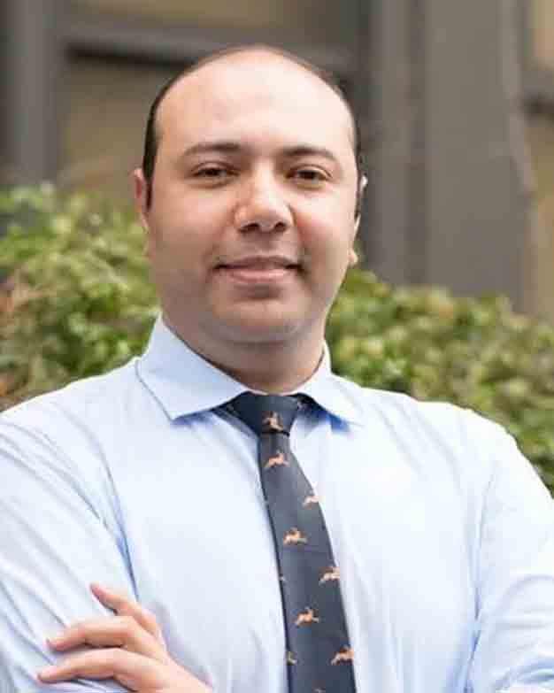Man with crossed arms wearing a blue shirt and patterned tie.