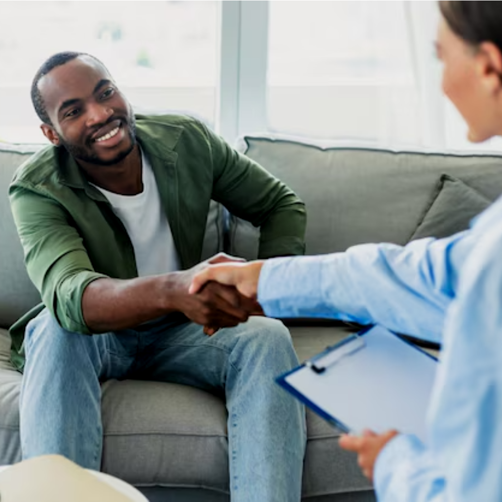 Man in green shirt shakes hands with a person holding a clipboard, sitting on a couch.
