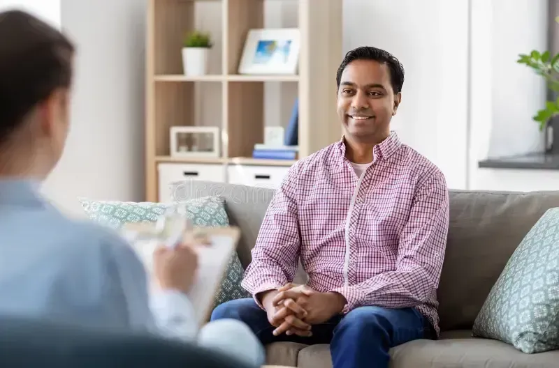 Man seated on couch, smiling at person holding clipboard; indoor setting.