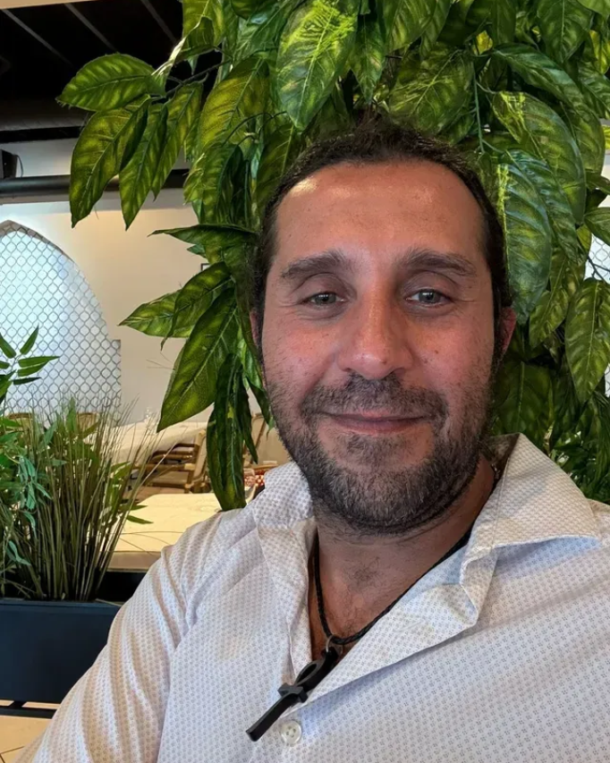 Man with short dark hair and a beard, smiling, wearing a white shirt, in front of leafy plants.