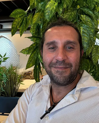 Man with short dark hair and a beard, smiling, wearing a white shirt, in front of leafy plants.