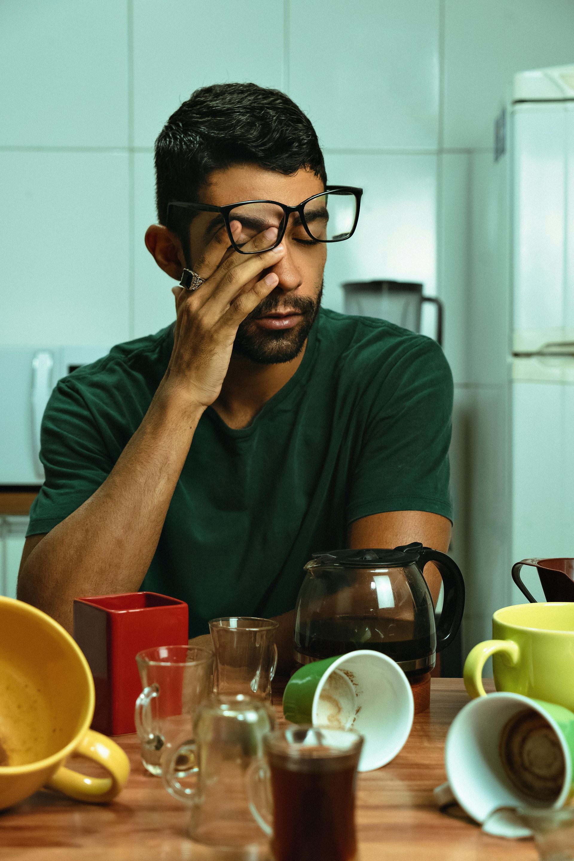 A person wearing glasses sits in a kitchen, looking tired with their hand over their eyes surrounded by empty coffee mugs.