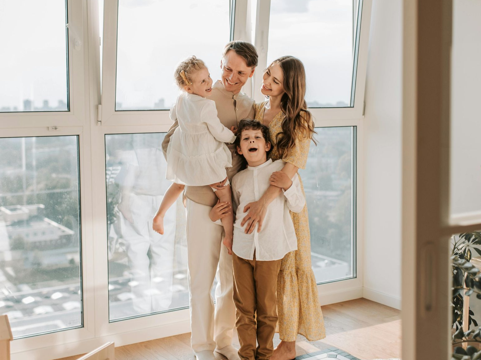 Family of four smiling by a large window; parents holding their children indoors.
