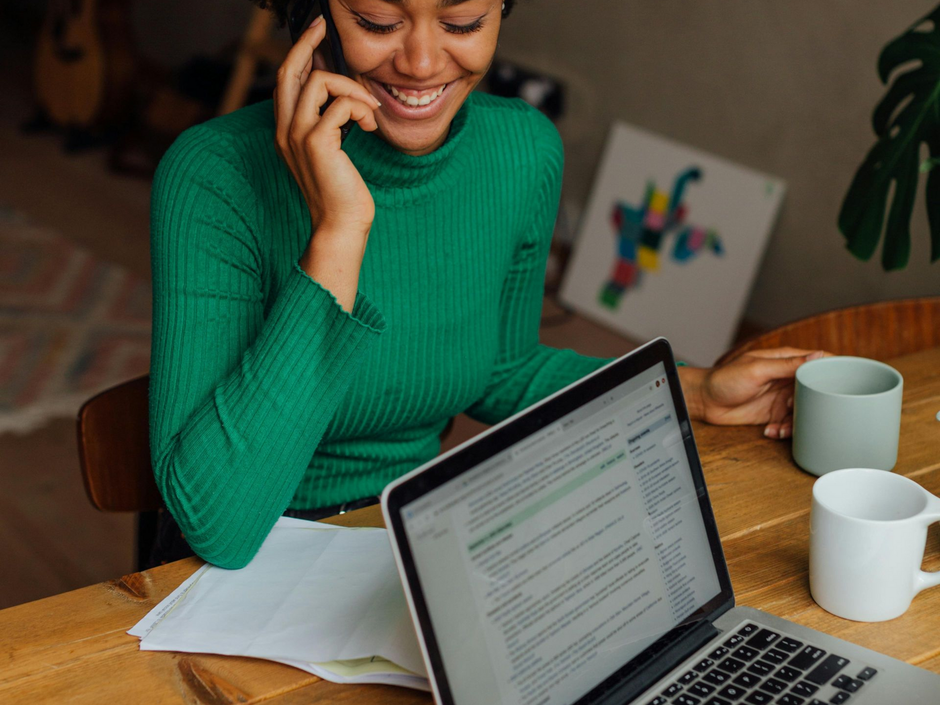 Woman in green sweater smiles while on a phone call, using a laptop, and holding a mug at a wooden table.