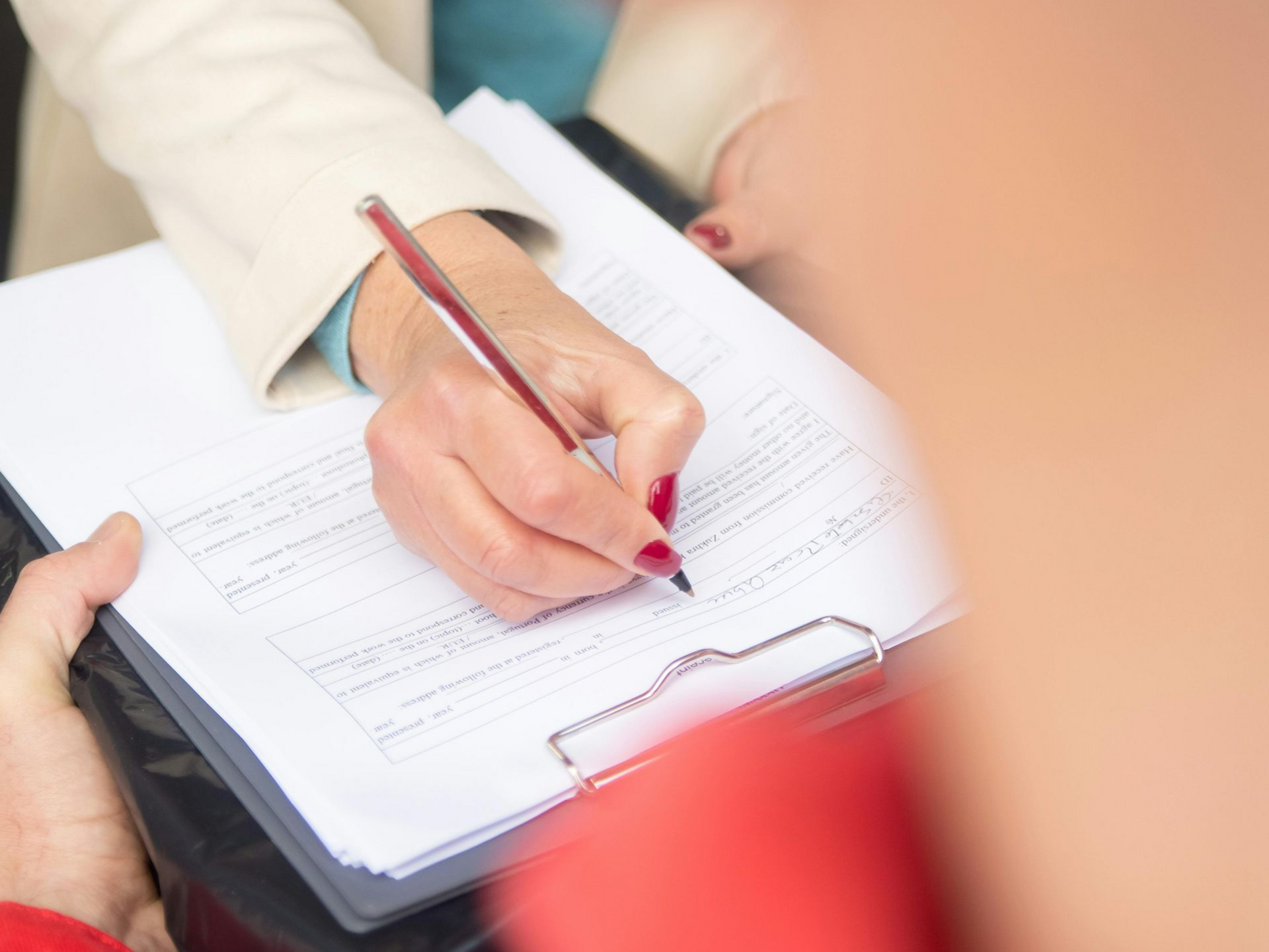 Person writing on a clipboard, possibly filling out a form, with another person's arm in the frame.