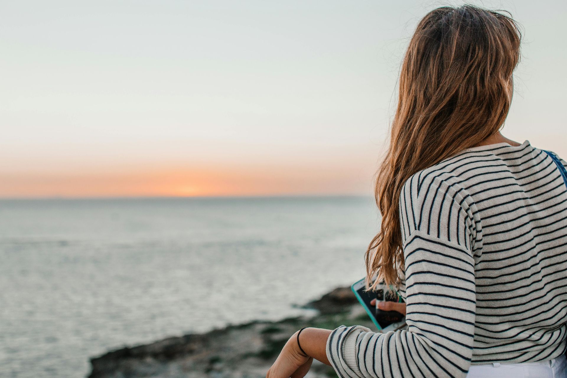 Woman with long hair and striped shirt overlooking ocean at sunset.
