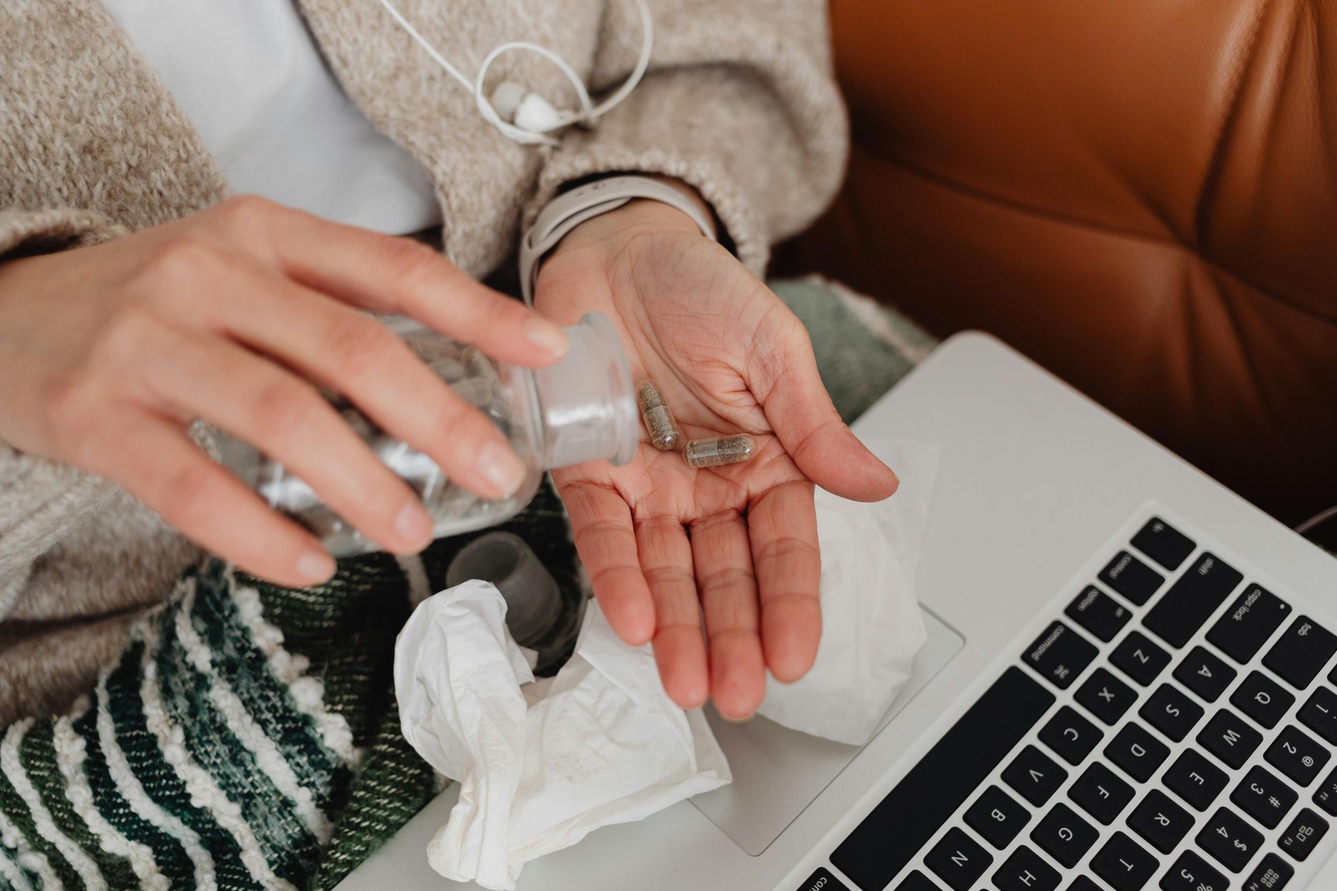Person pouring pills from a bottle into their hand next to a laptop and tissue.