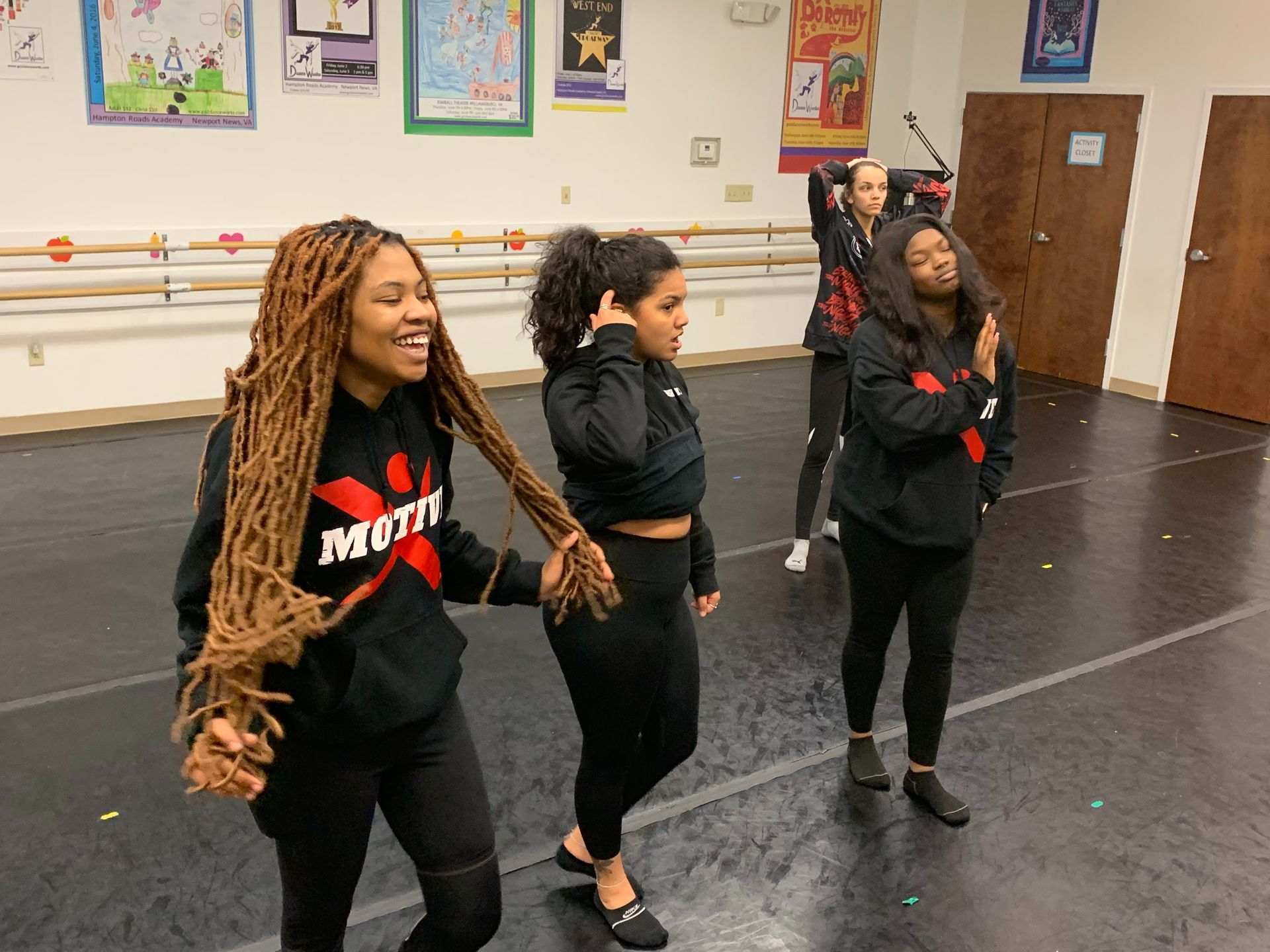 Four people in a dance studio wearing black hoodies and leggings. One holds another's long hair, smiling.