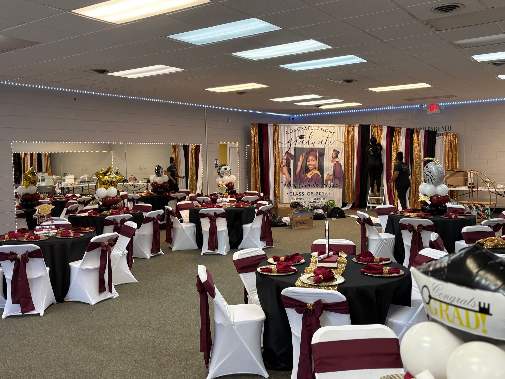 Event hall with tables decorated in black, white, and burgundy. Backdrop with a photo and gold and black drapes.
