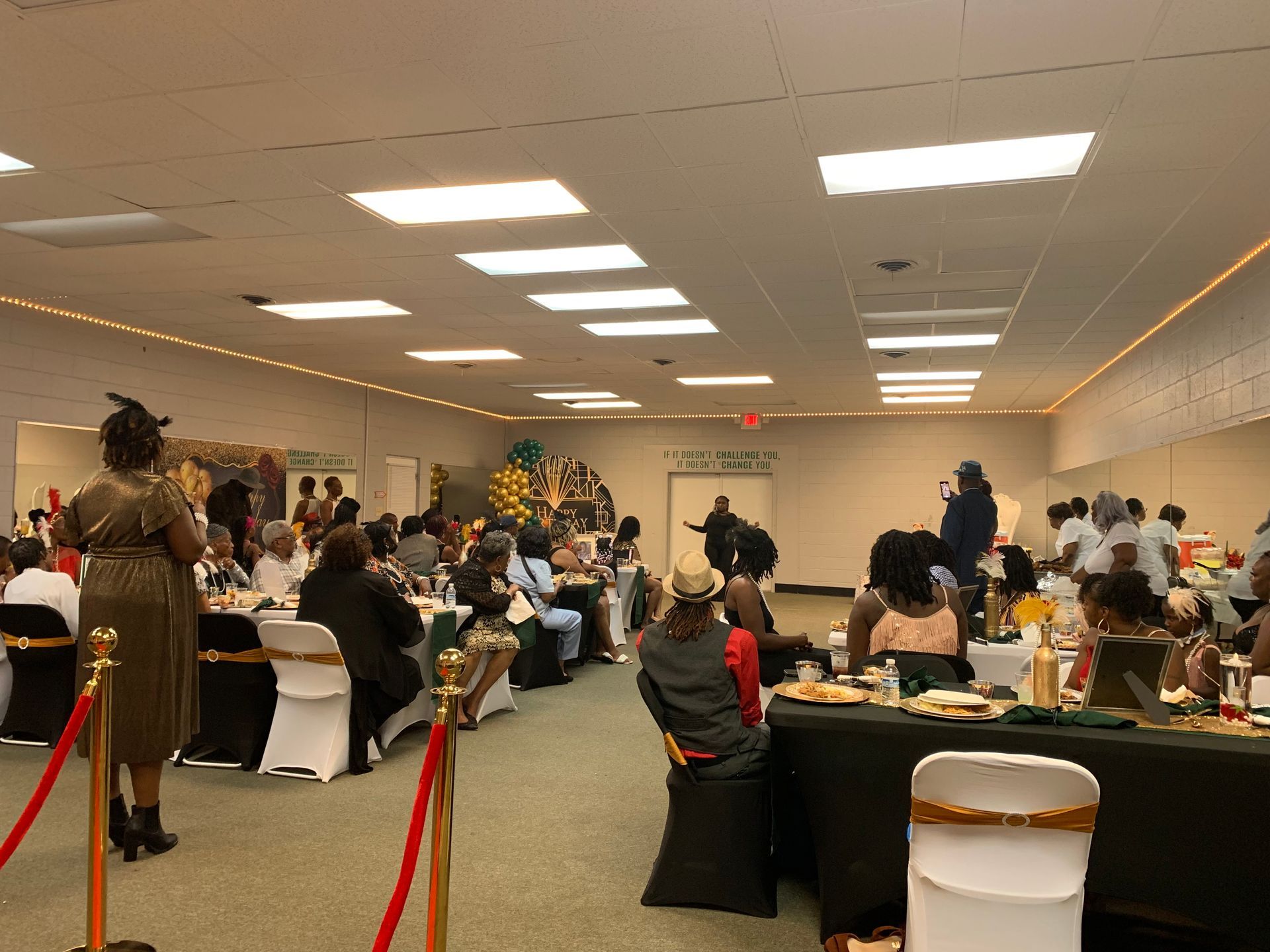 Event in XMotive Dance Studio, people seated at tables, a person speaking from a stage, red rope barriers.