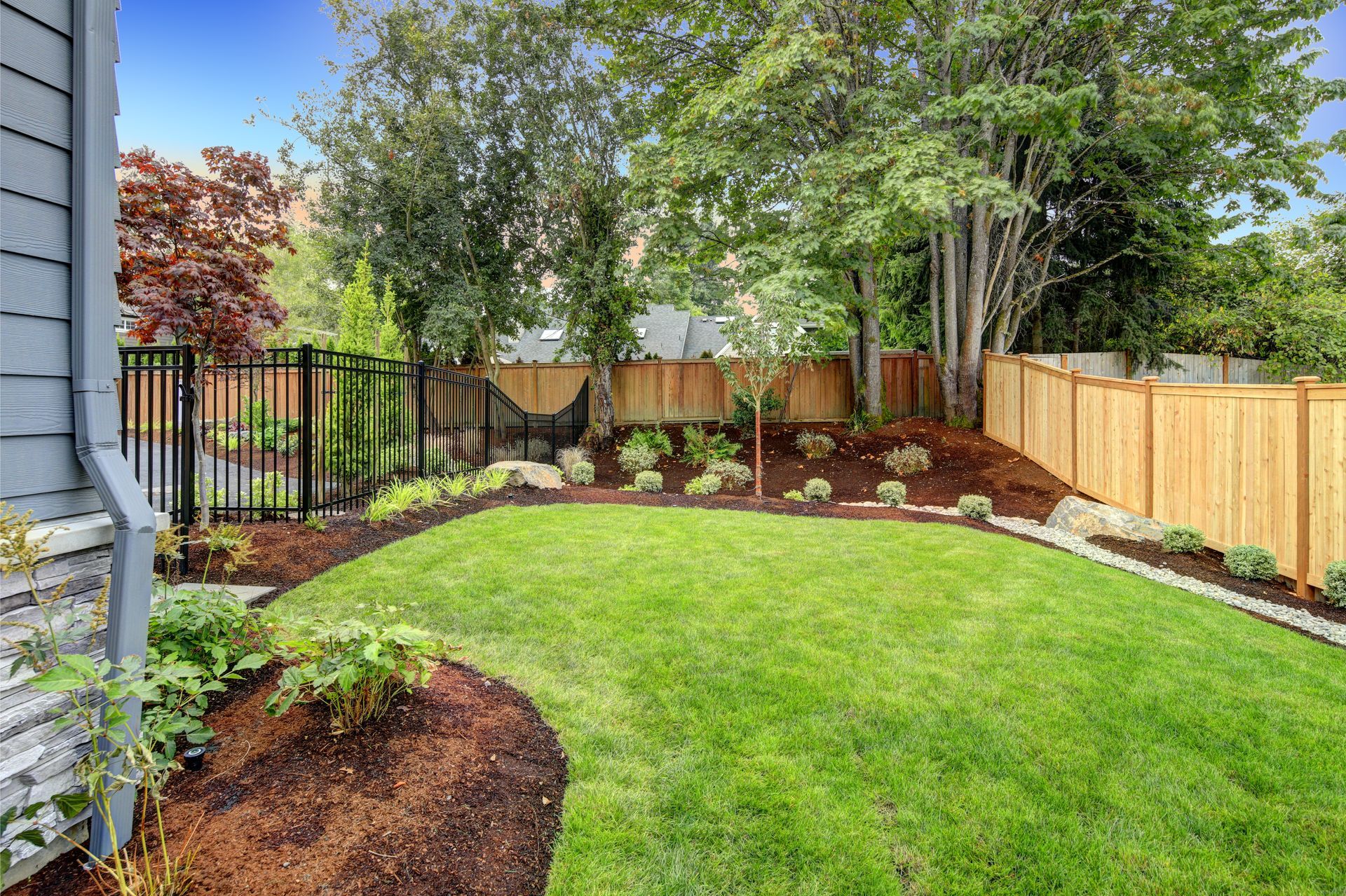 Lush green backyard with lawn, flower beds, wooden fence, and tall trees along the edge