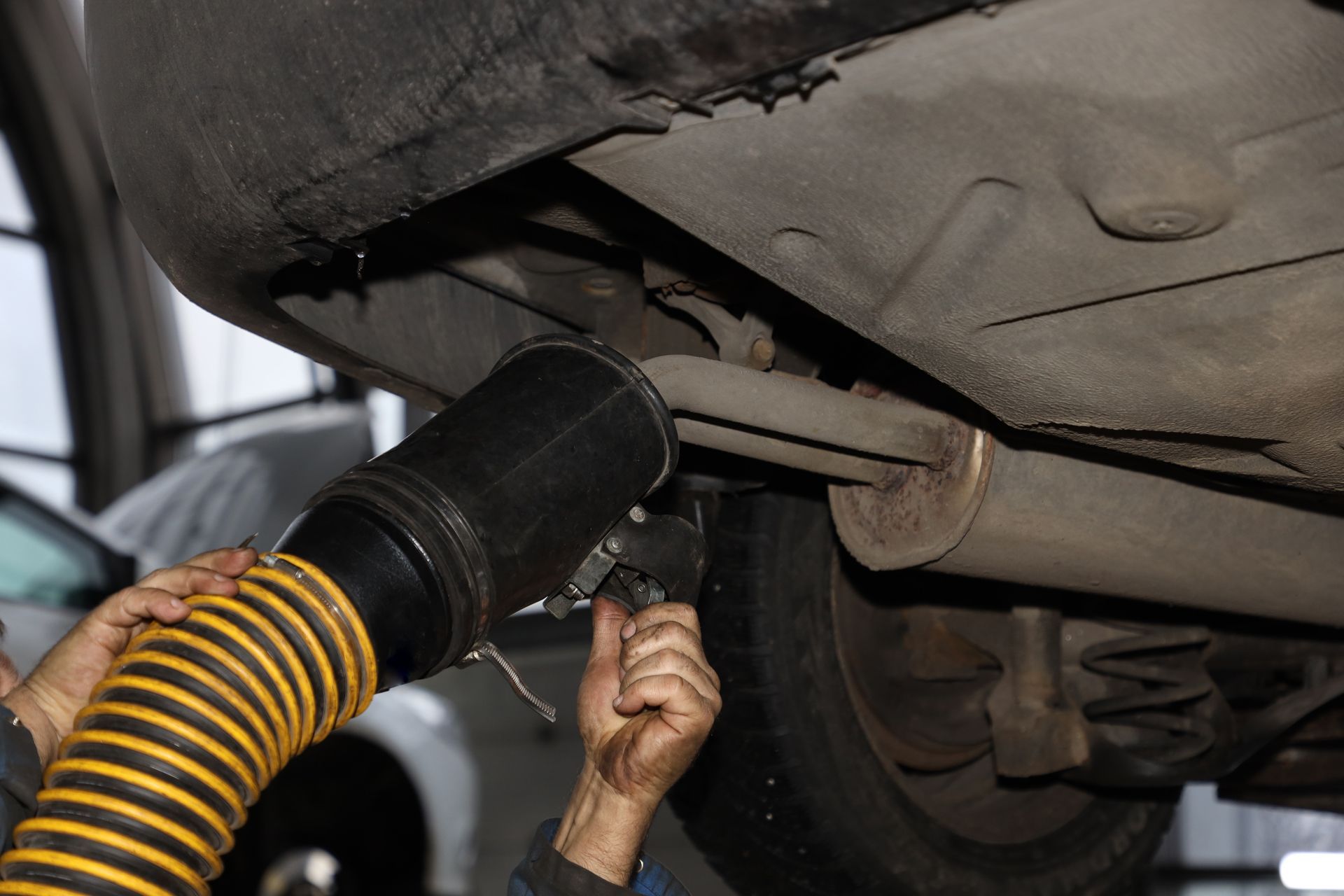 Mechanic working on the underside of a car, using a hose to connect to the exhaust system.