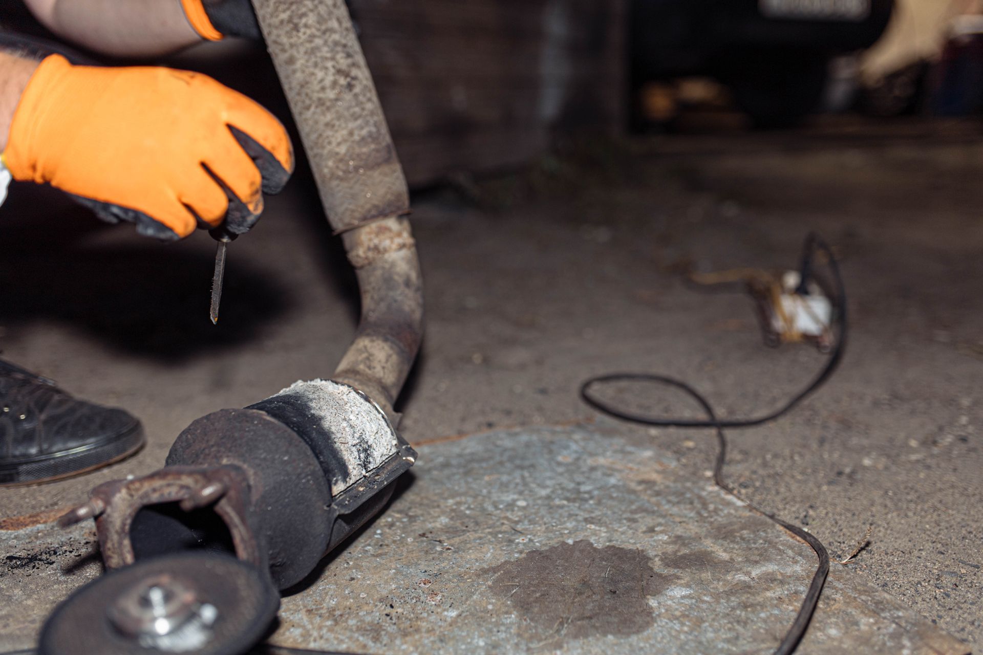 Person wearing orange gloves cutting a car exhaust pipe with a tool in a garage.