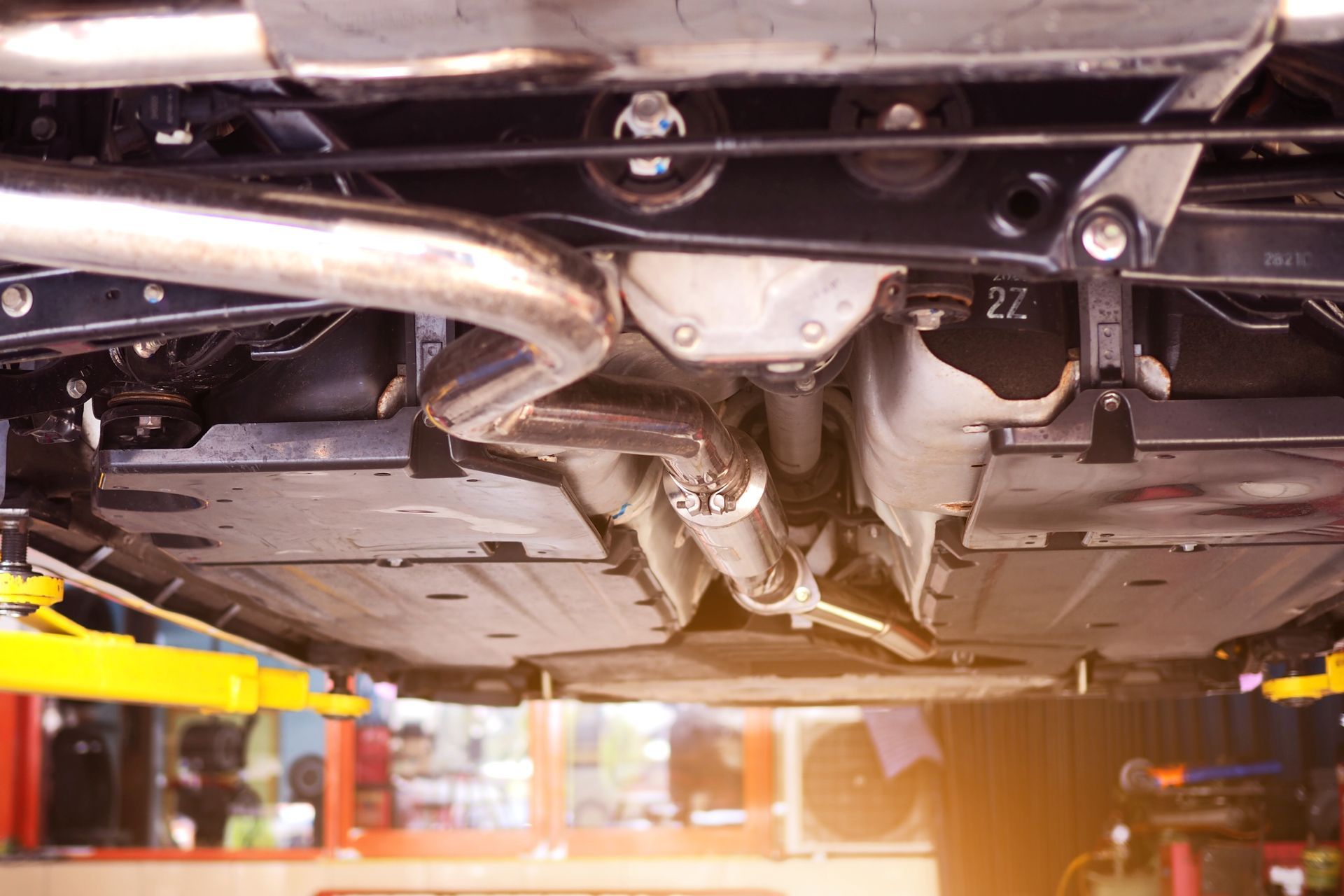 Underside of a car on a lift in a repair shop, showing exhaust system, metal components, and protective paneling.