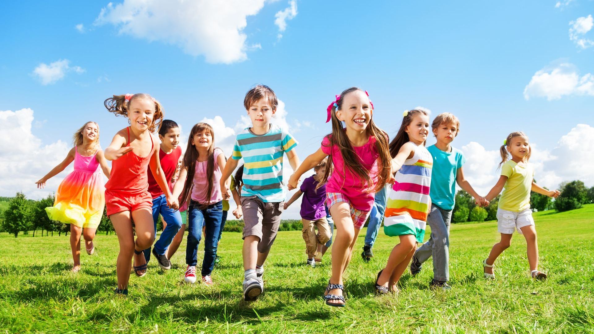 Children running on a green grassy field under a blue sky, smiling and holding hands.