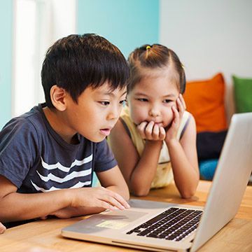 A boy and a girl are sitting at a table looking at a laptop computer.