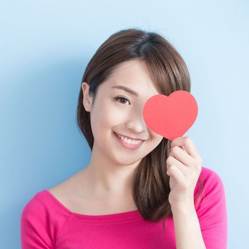 A woman in a pink shirt is holding a red heart in front of her eye.