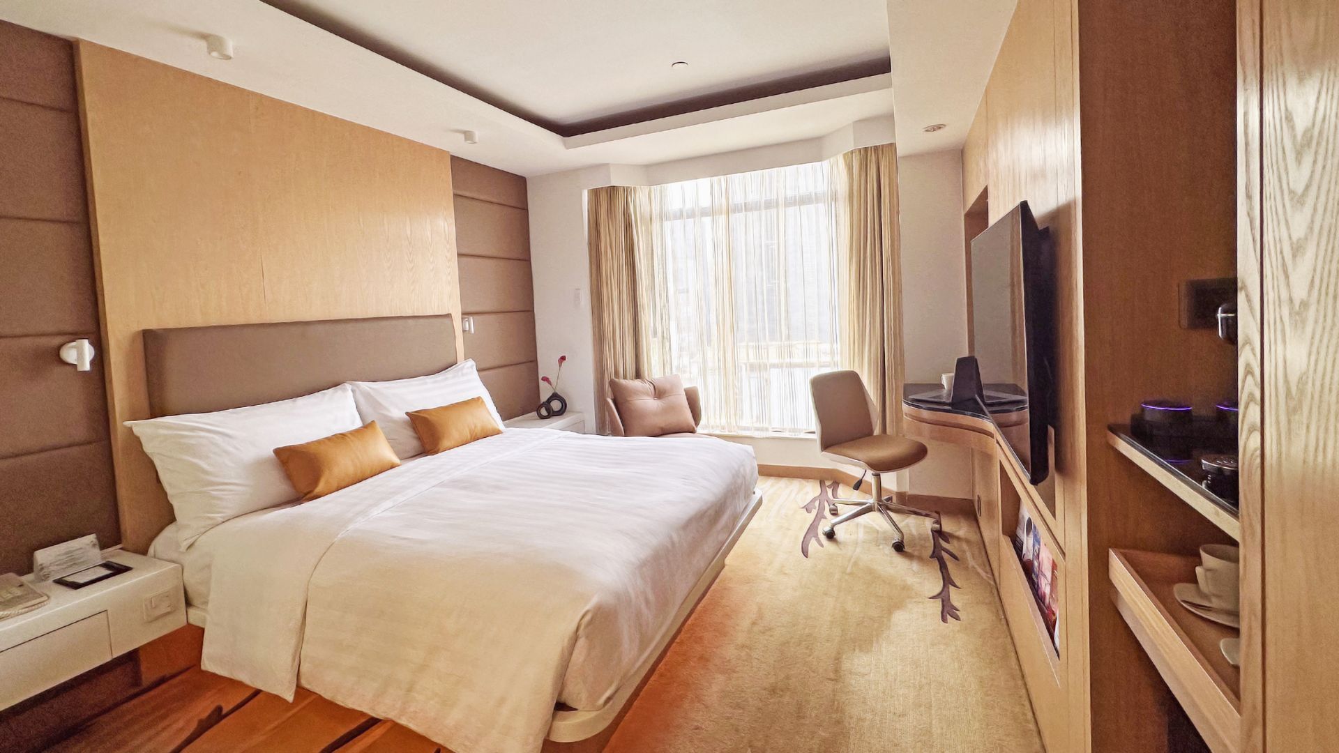 Hotel room with a bed, desk, and window with a city view.  Decorated in warm tones with a modern design.