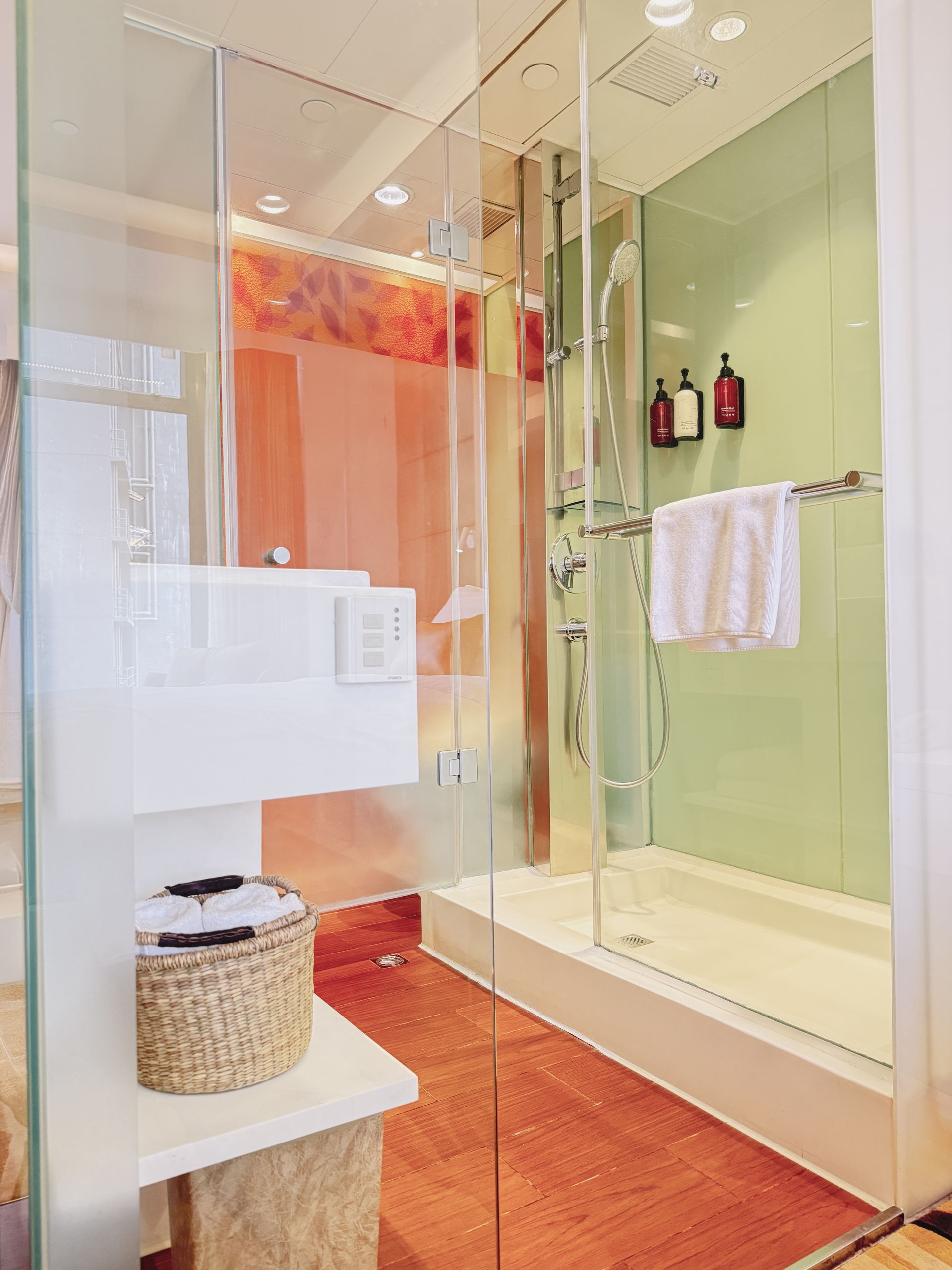 A modern bathroom with a glass-enclosed shower, light green walls, and an orange wooden floor.  A white towel hangs near shower toiletries.