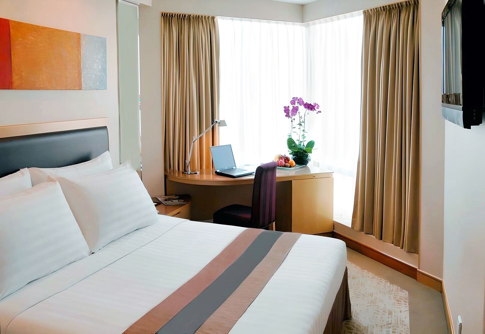 Bedroom interior with a bed, desk with laptop, window with curtains, and a decorative flower arrangement.
