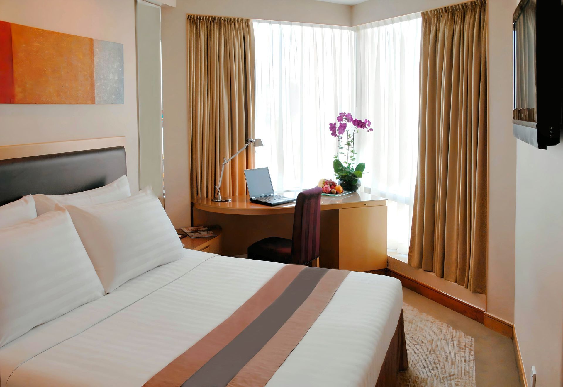 A hotel room with two twin beds, a desk by a window, and a floral painting. The room is beige and has a lamp on a nightstand.