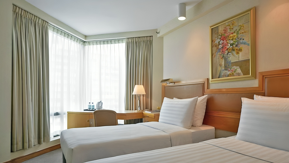 A hotel room with a bed, chair, and a window with curtains. A small couch is visible in the adjacent room.