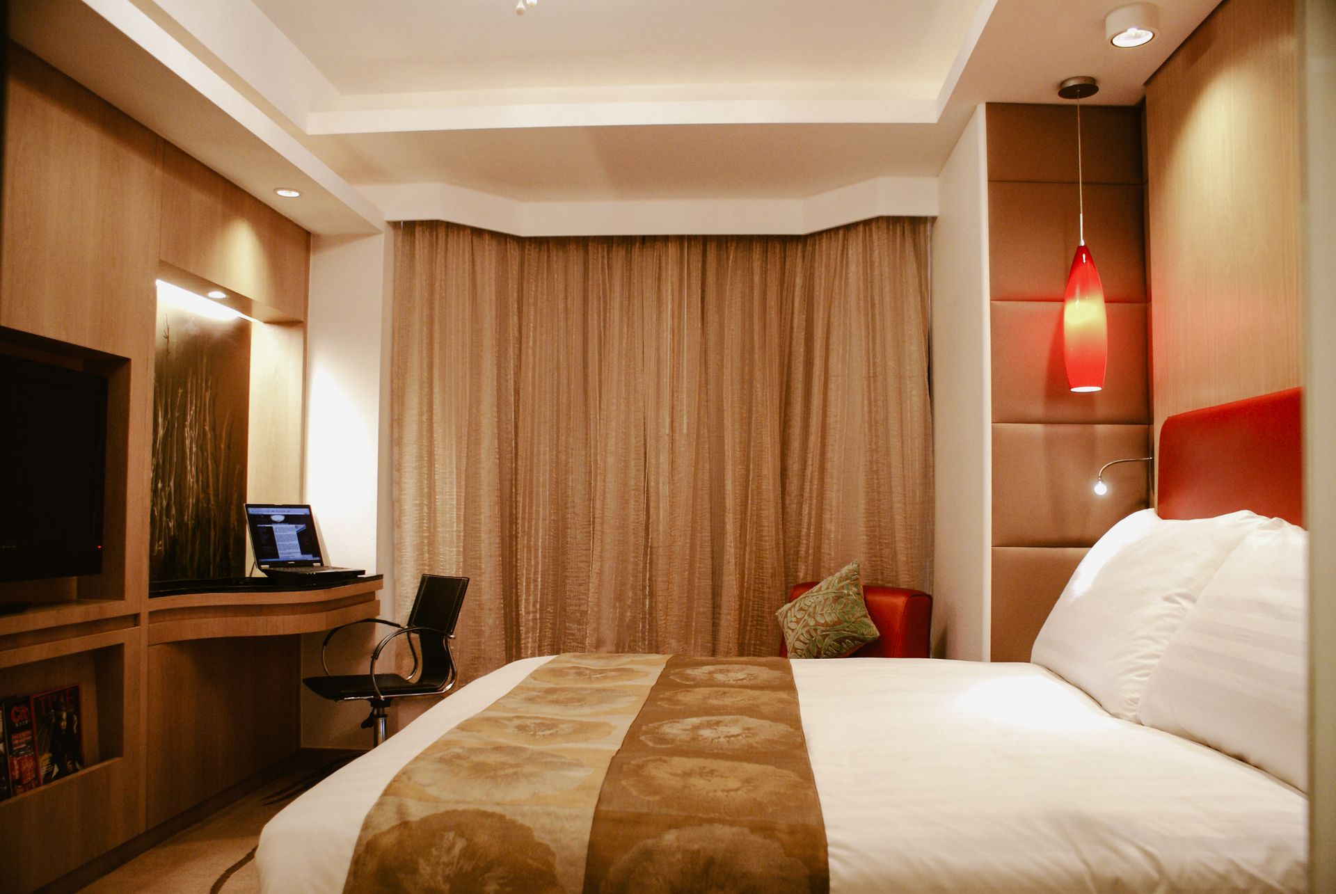A hotel room with a bed, desk, and TV. The room is decorated in warm colors with a red accent light.