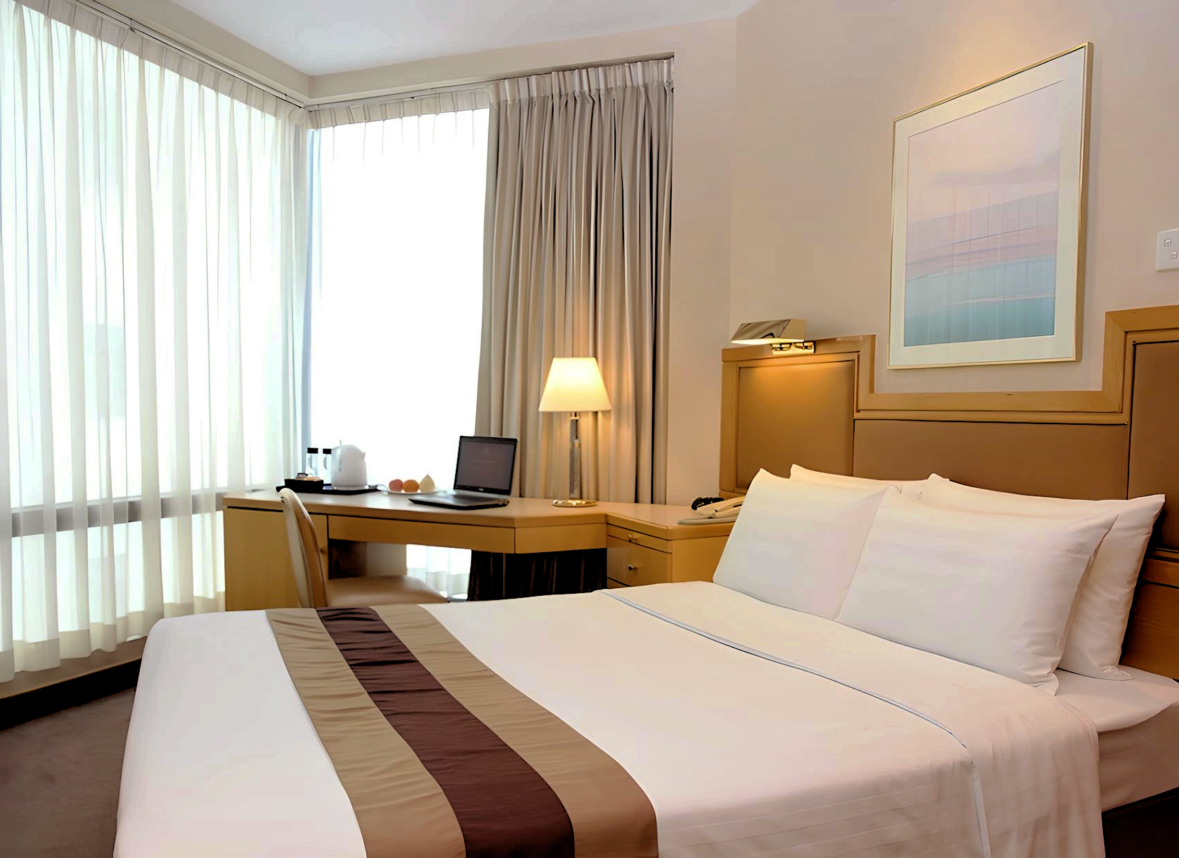 Hotel room with a bed, desk, window, and lamp. The bed has white sheets with a brown stripe. A laptop sits on the desk.