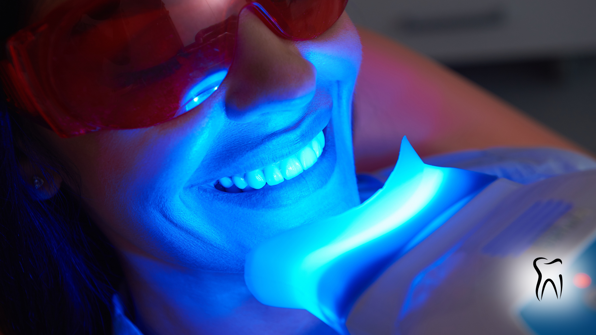 Person undergoing teeth whitening treatment with protective glasses, illuminated by blue light.
