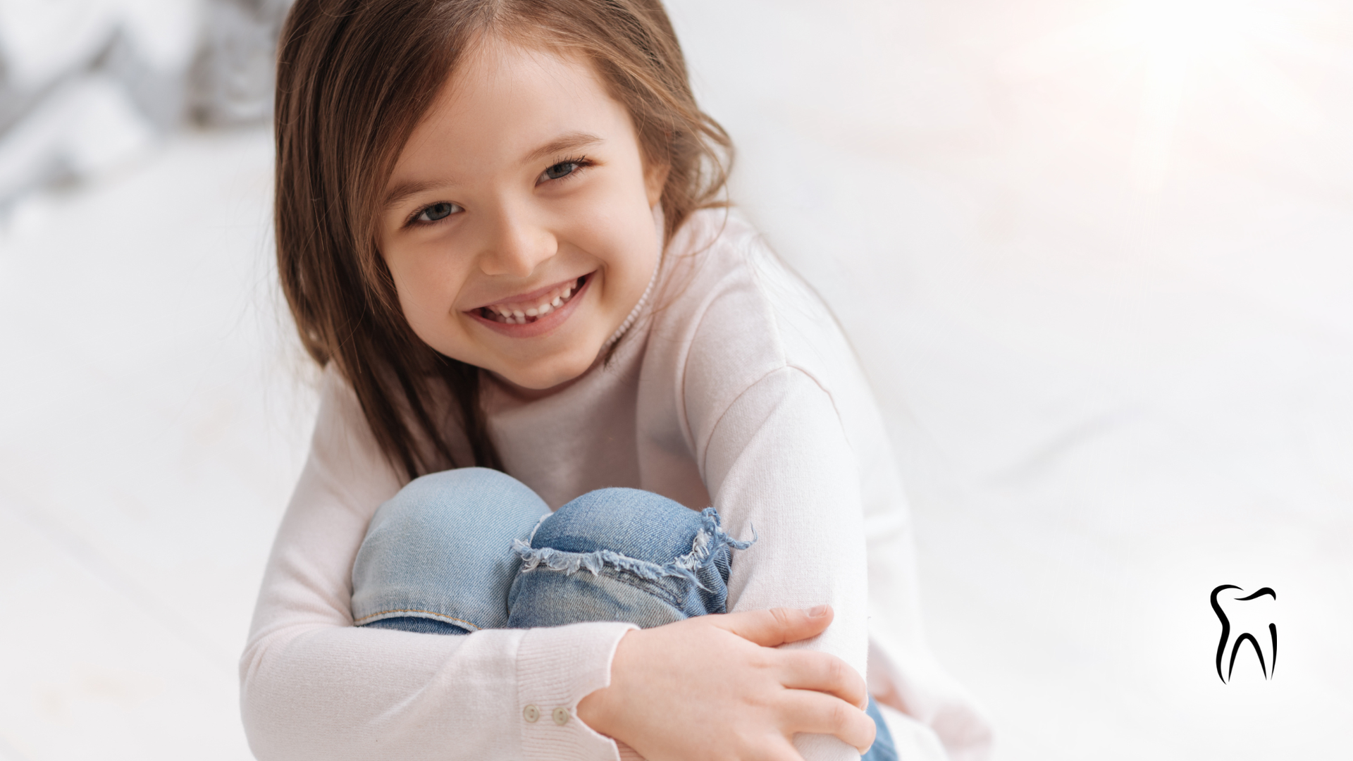 Young child smiling, seated with knees up, wearing light top and blue jeans, dental icon in the corner.