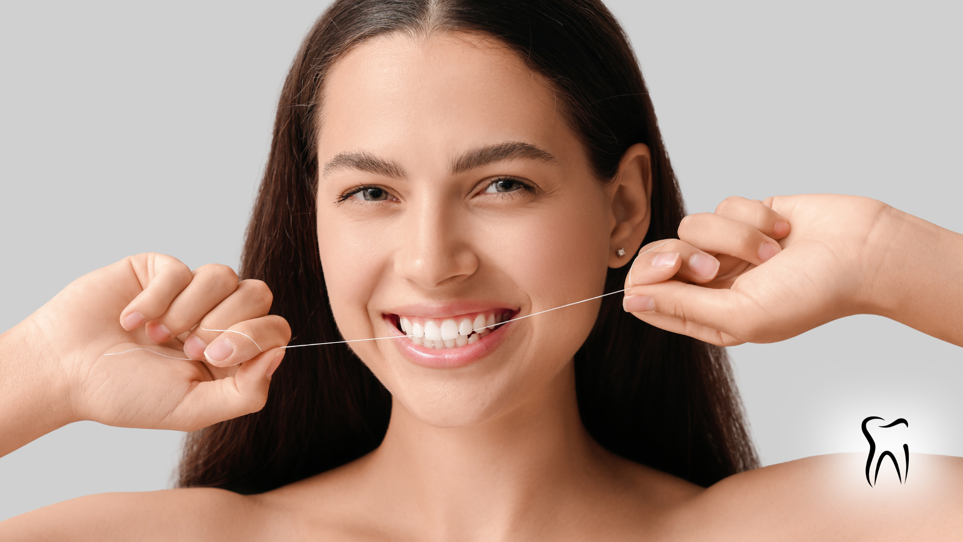 Woman flossing teeth, smiling at camera against a gray background.