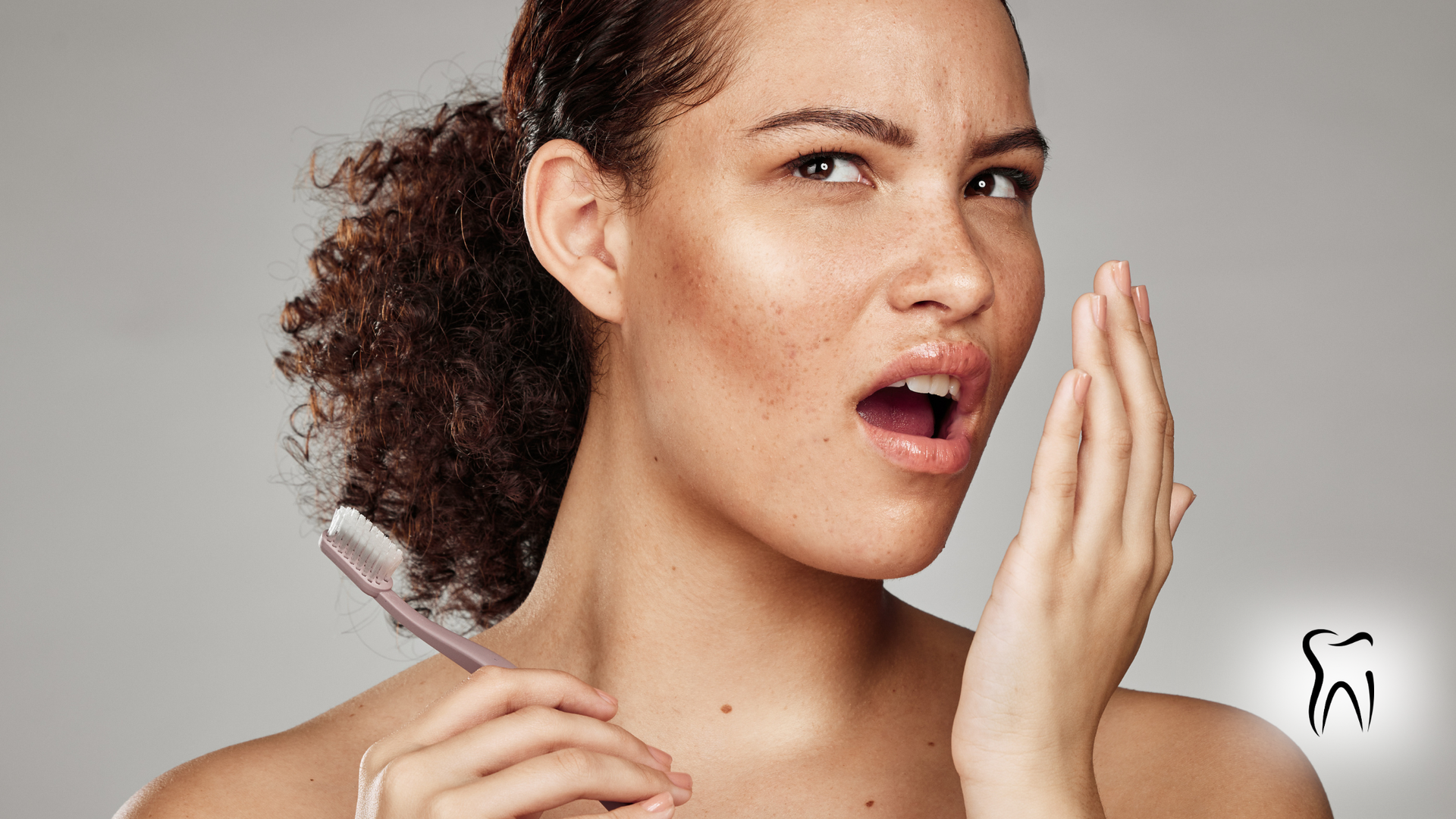 Woman holding toothbrush, checking breath with hand. Open mouth, worried expression.