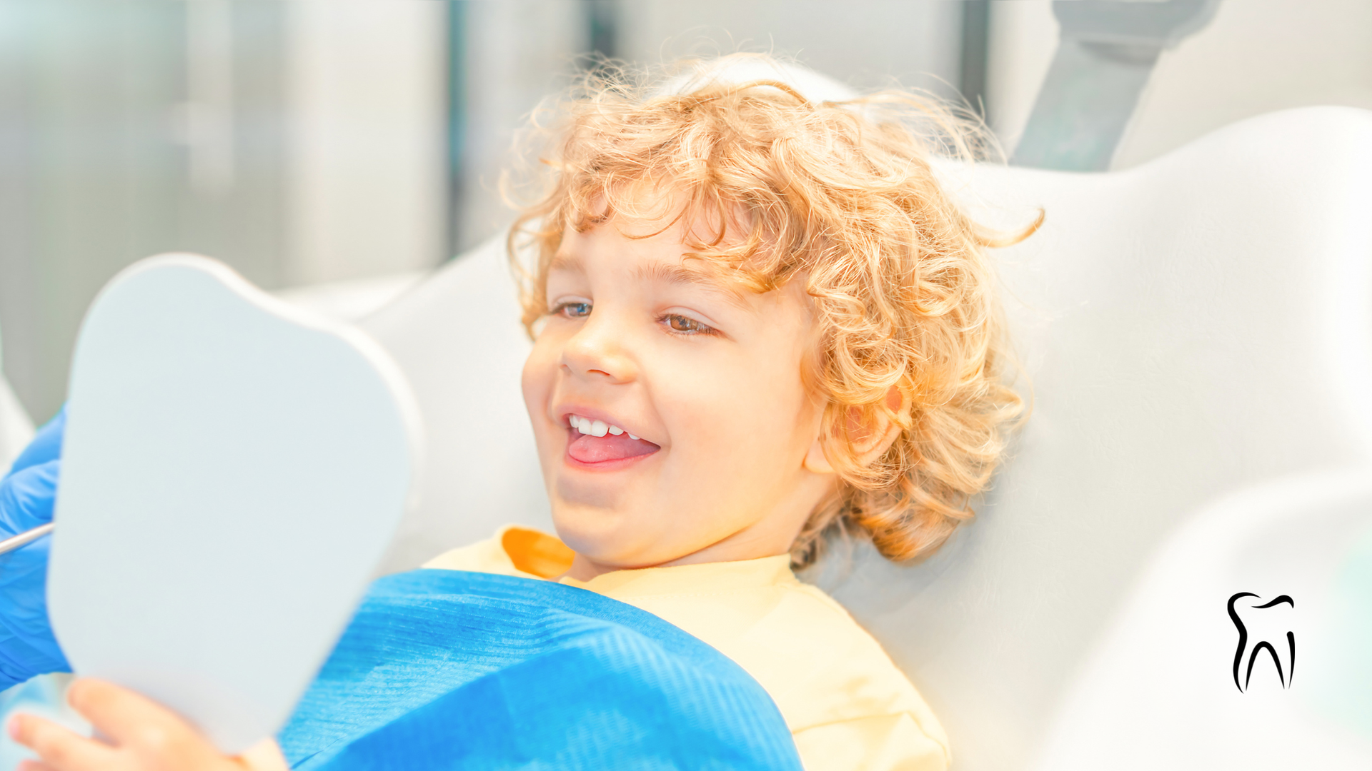 Boy with curly hair smiles while looking at teeth in a mirror at the dentist.