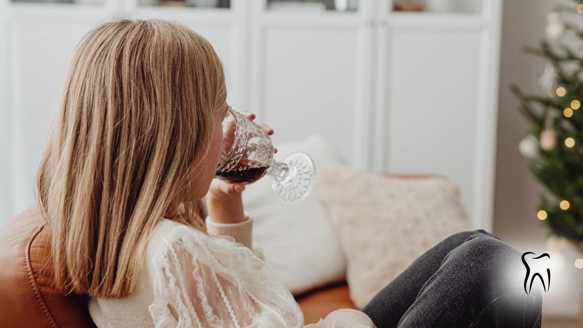 Woman drinking red liquid from a glass, sitting near Christmas tree.