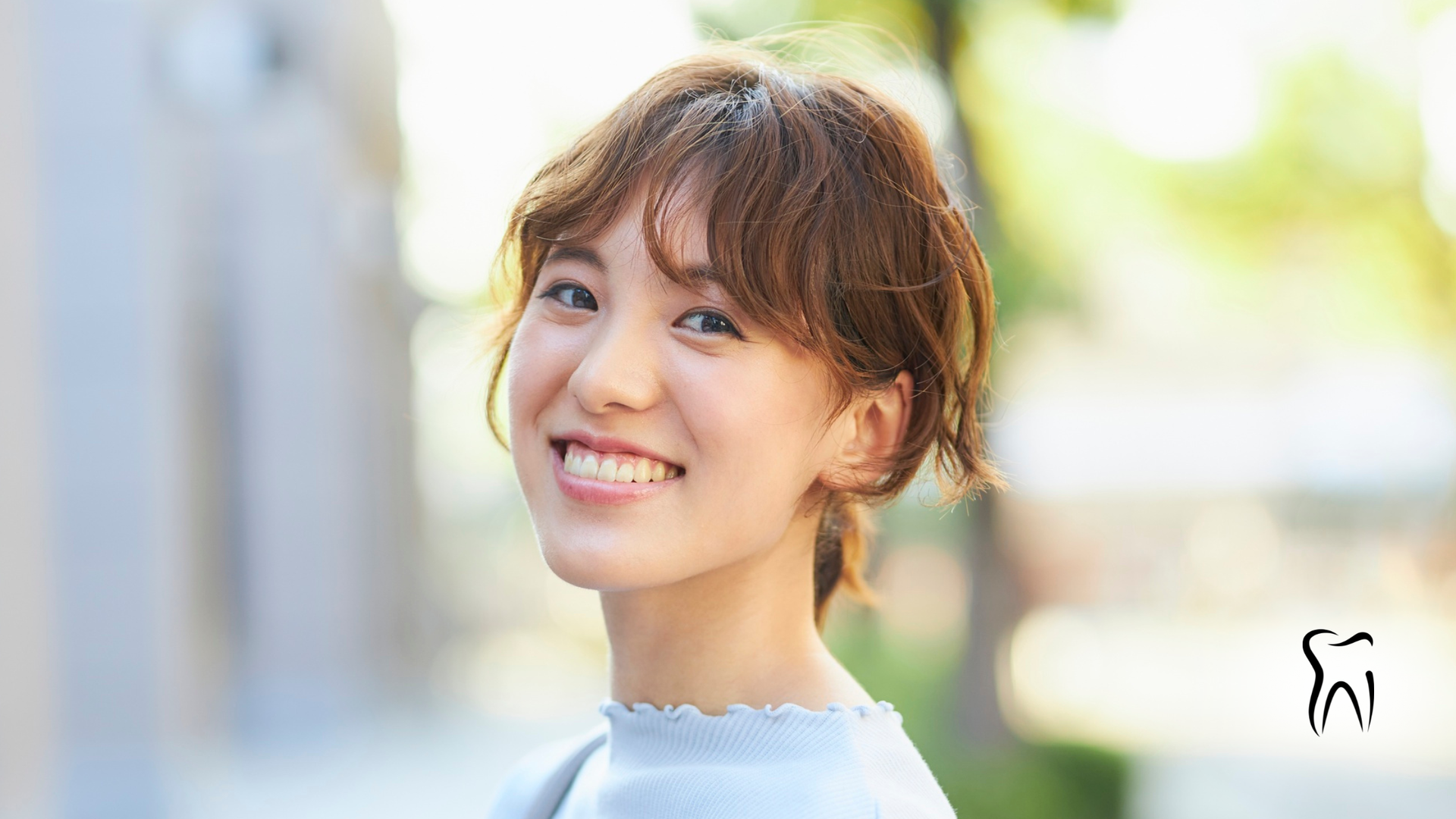 Woman smiling with teeth exposed, short brown hair, light blue shirt, outdoor setting.
