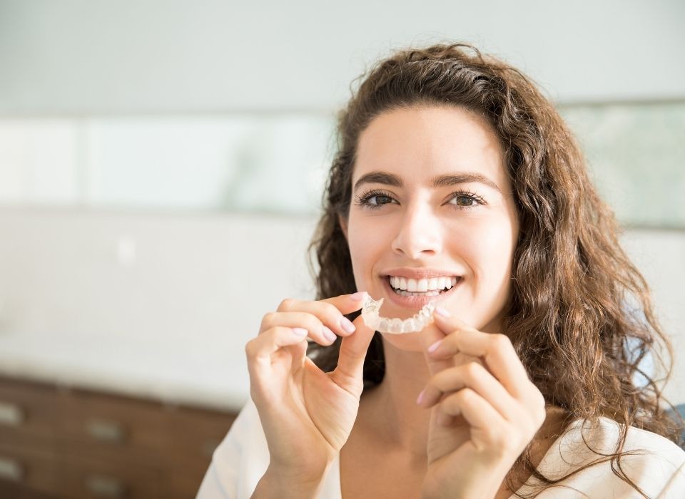 Woman holding clear aligner, smiling. Indoor setting.