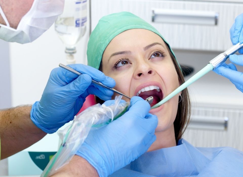 Woman in dentist chair, mouth open. Dentists work in mouth with tools, wearing blue gloves.