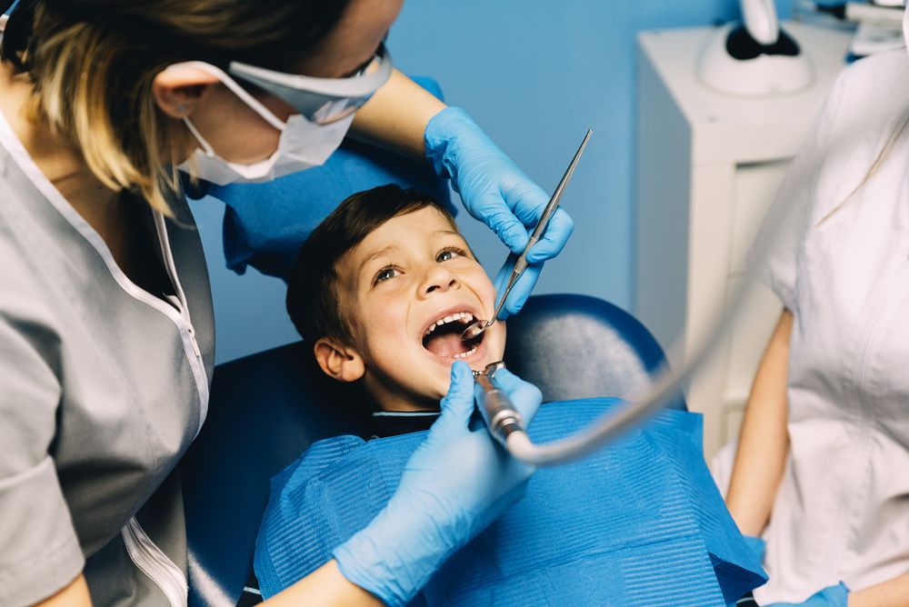 Boy at dentist with mouth open, dentist examining teeth with tools. Blue and white color scheme.