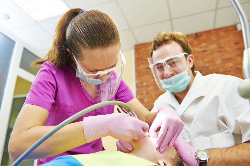 Dentist and assistant examining a patient's mouth; they wear masks and shields in an office setting.