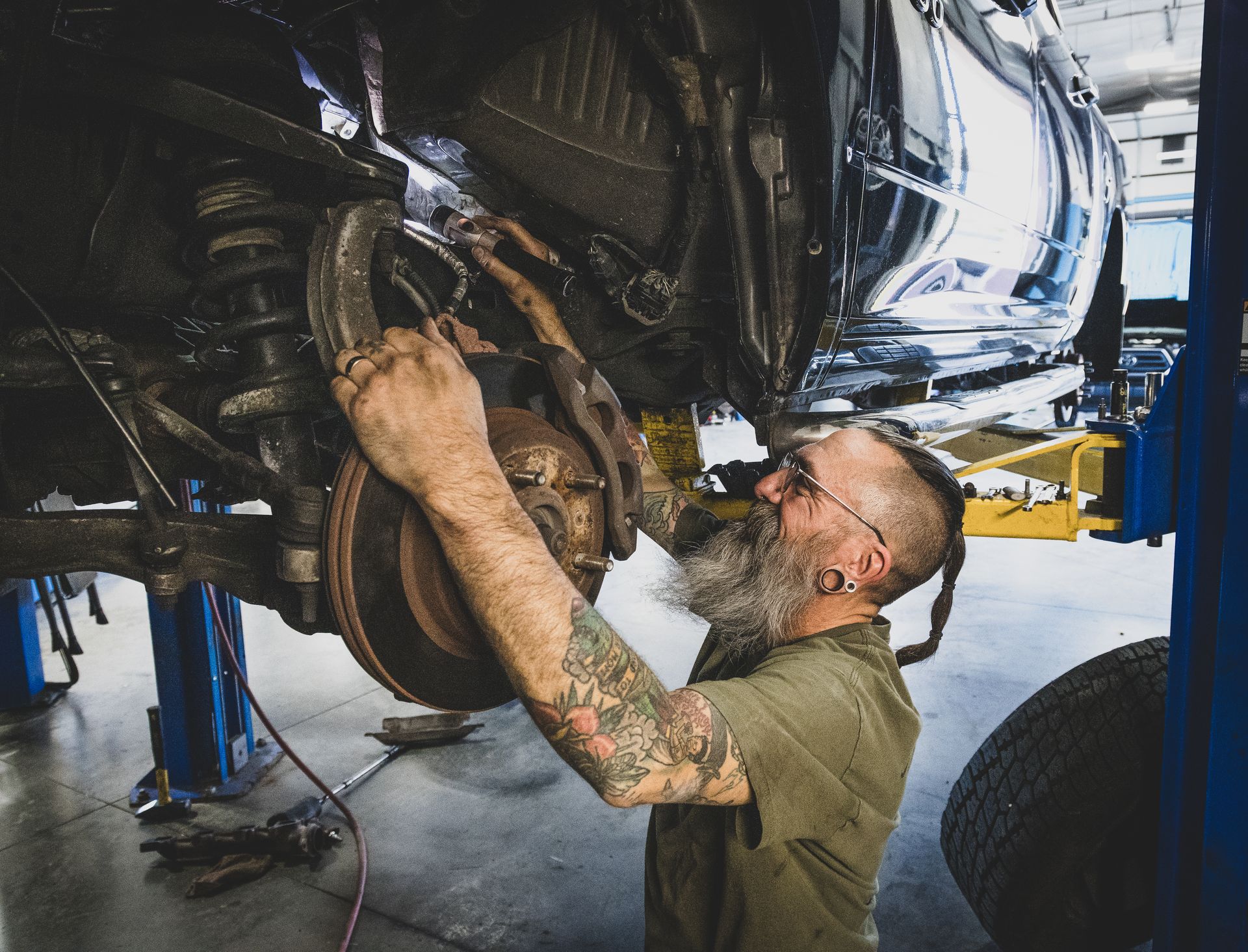 Mechanic working on car's suspension. He has tattoos and a beard, in a garage setting with a car on a lift. | Mountain Top Service Center