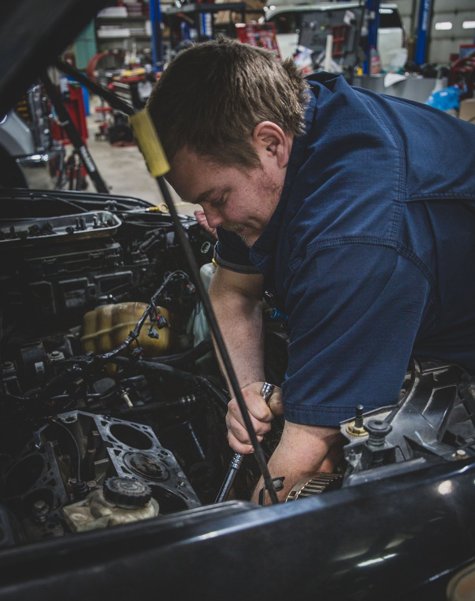 Mechanic in a blue shirt working on a car engine in a garage; he is focused on his task. | Mountain Top Service Center