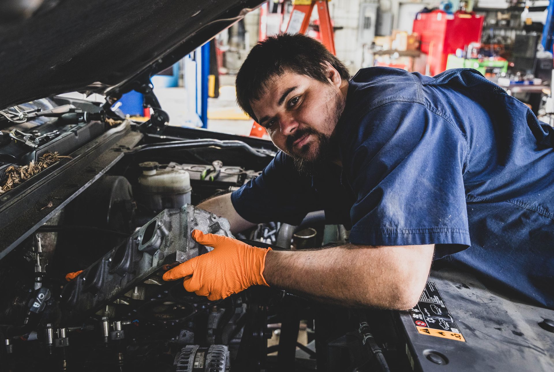 Mechanic working on car engine, wearing orange gloves and blue uniform. Garage setting. | Mountain Top Service Center