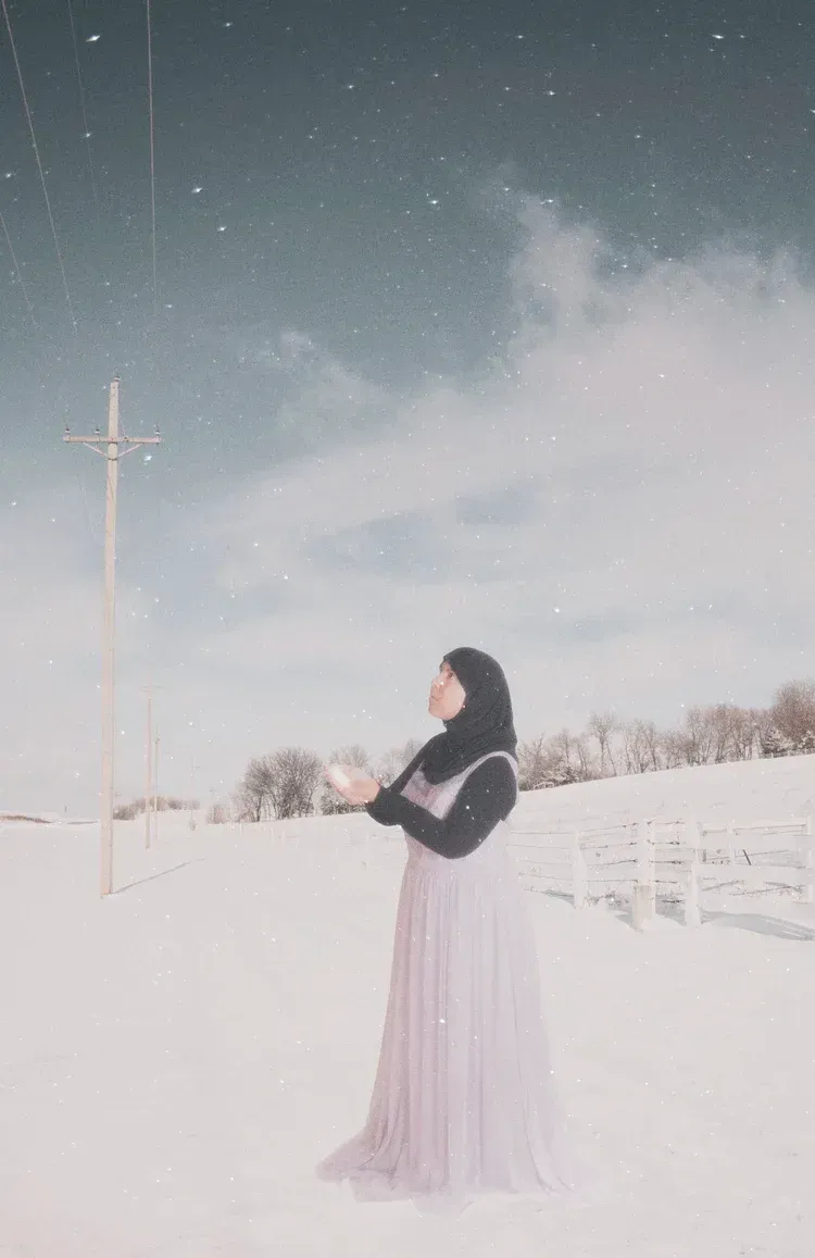 Woman in a long, lavender dress and hijab stands in a snowy field, arms outstretched, under a starry sky.