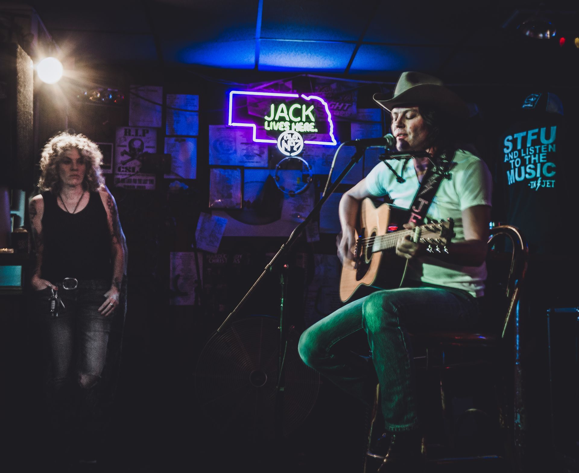 Man in cowboy hat plays acoustic guitar on stage, another person stands next to him at a dimly lit bar.