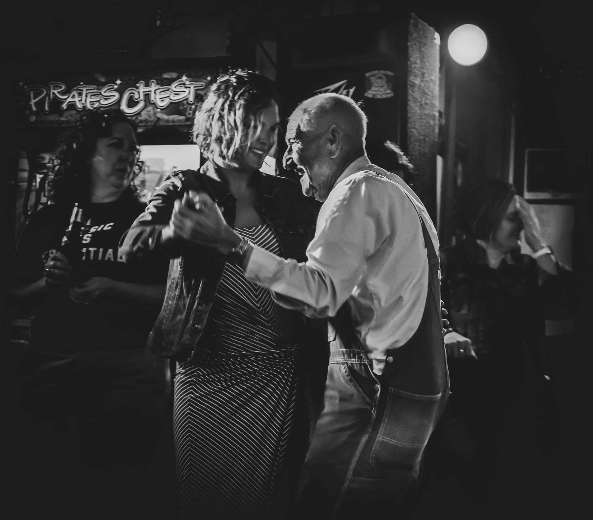 Elderly man dances with a woman in a dimly lit bar, others watch.